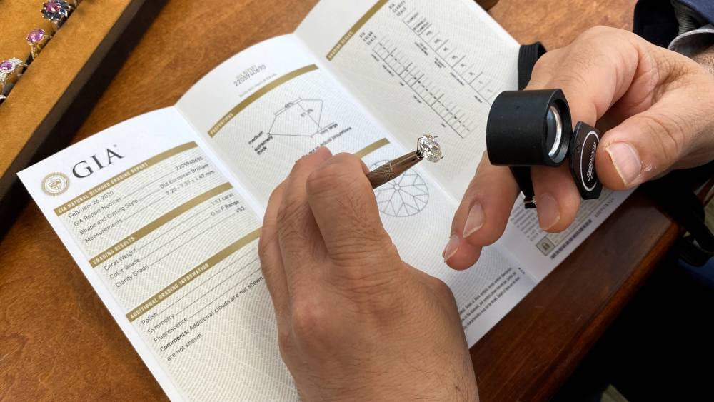 An open GIA grading report lying on a wooden desk, one hand holding a loose round diamond in fine tweezers above the page and another hand holding a black loupe, with the proportions diagram and the round clarity plot printed on the open spread and a tray of rings at the edge of the desk