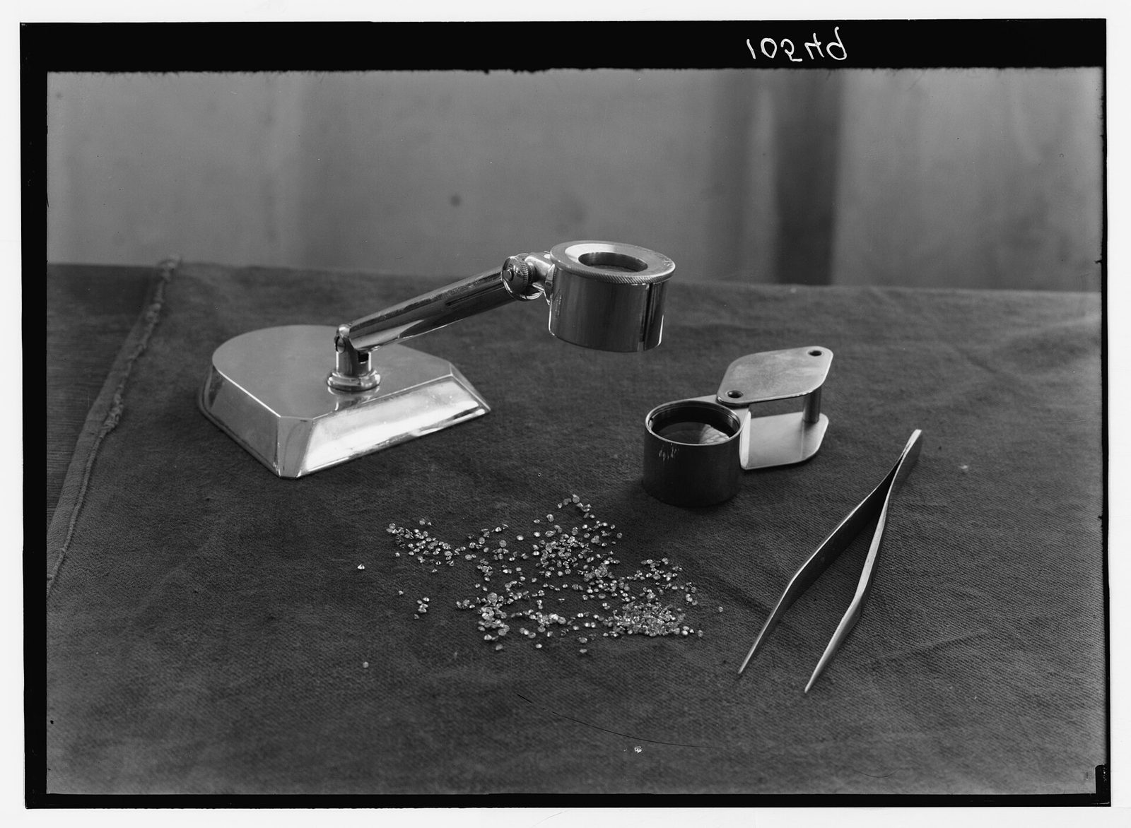 Archival black and white photograph of a jeweler's loupe stand, a folding loupe, tweezers and a small scattered parcel of polished diamonds on a cloth covered table