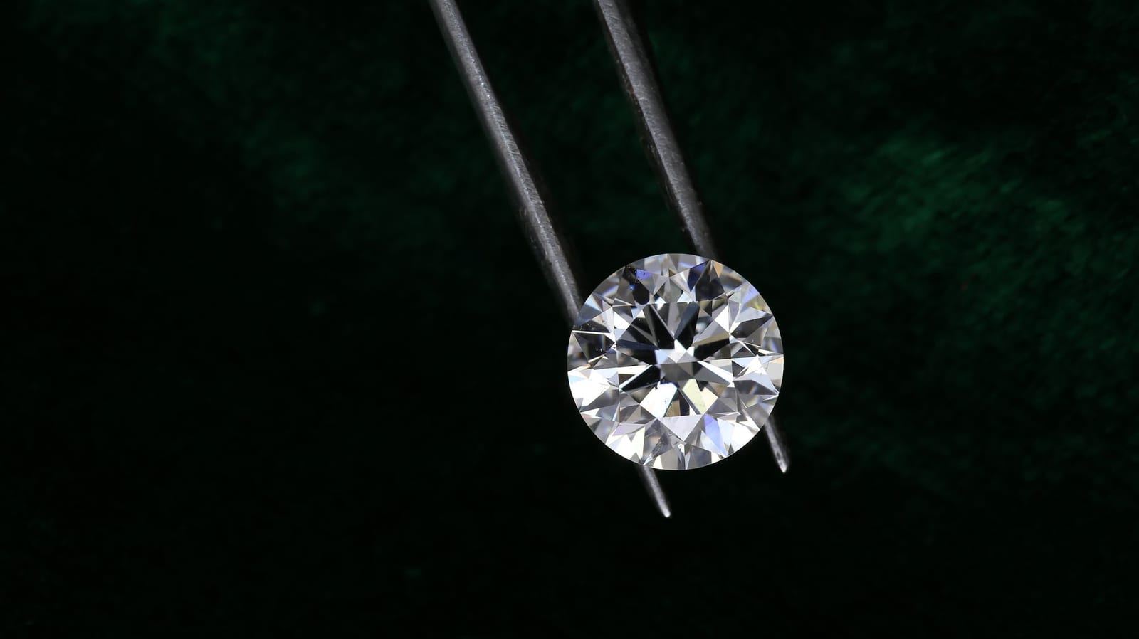 A round brilliant cut diamond held face up in a pair of steel stone tweezers, lit against dark green cloth so the table, the star facets and the girdle outline read clearly