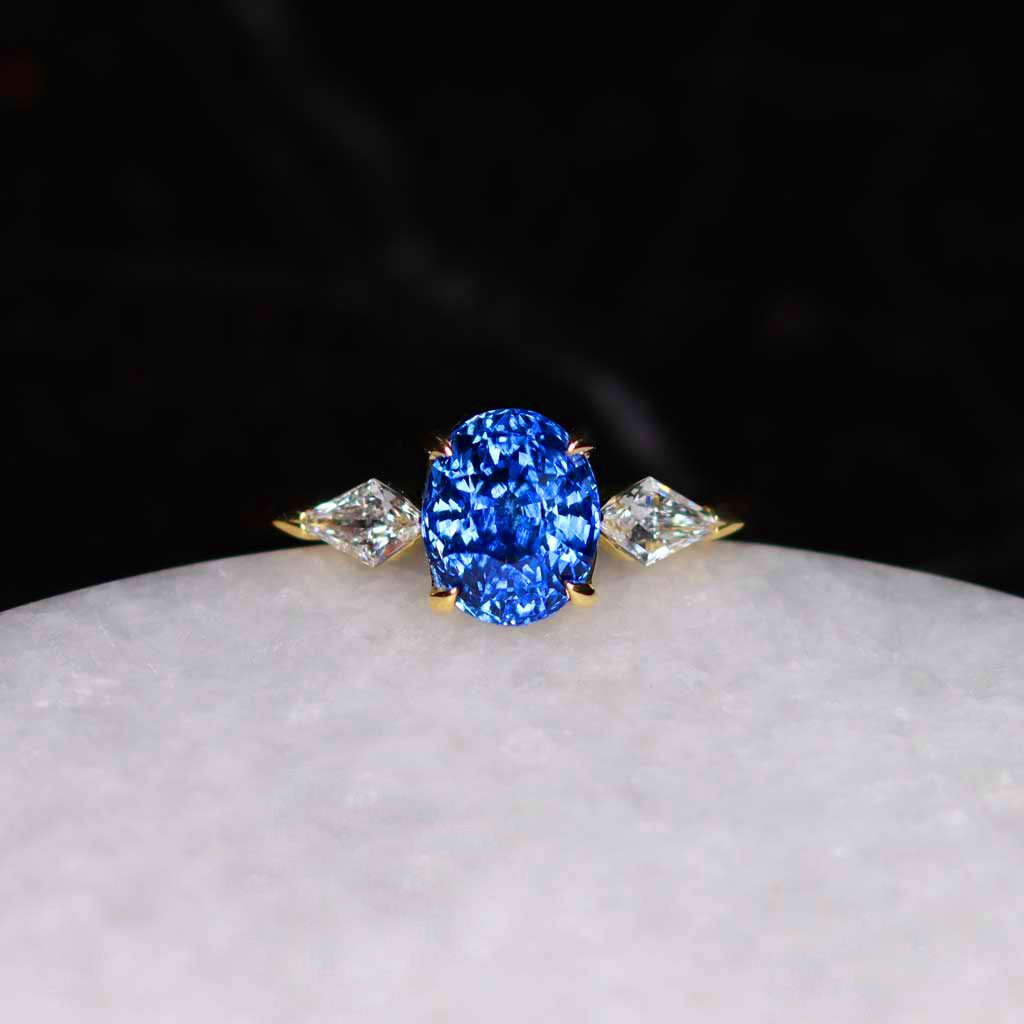 A three stone engagement ring with an oval blue center stone flanked by two kite shaped diamond side stones, set in yellow gold
