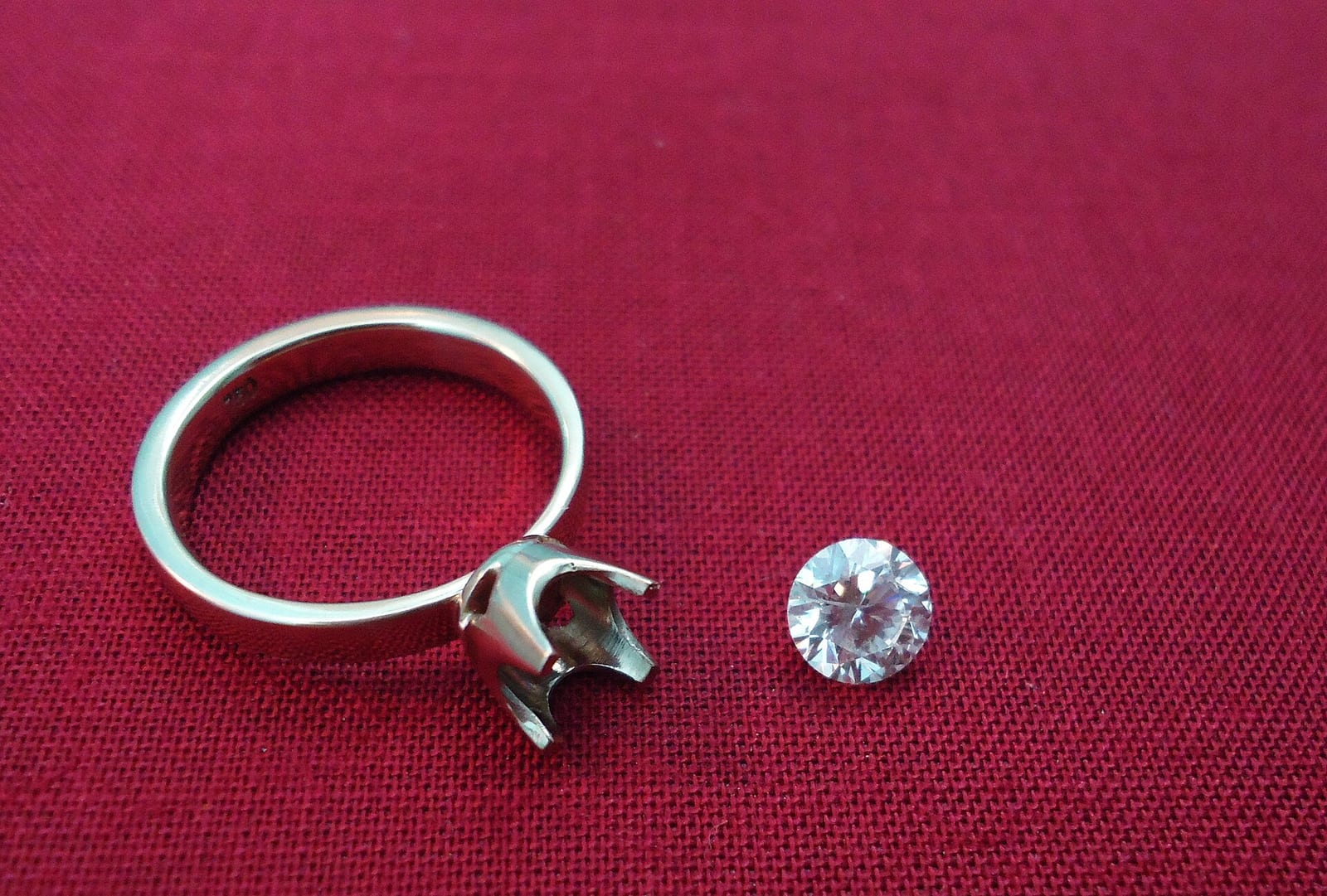 An empty white gold solitaire mounting with open prongs lying on red cloth next to a loose round brilliant diamond that has not been set yet