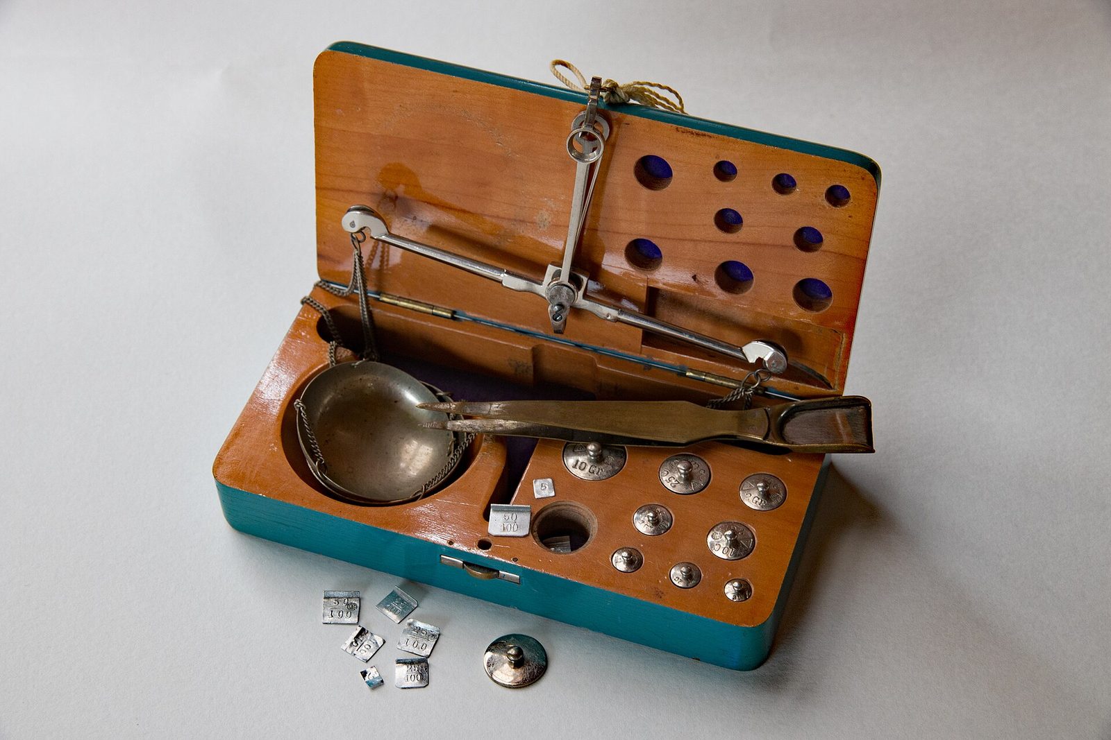 A vintage jeweler's balance scale open in its case, with small brass carat weights and a pair of tweezers laid out beside the weighing pan