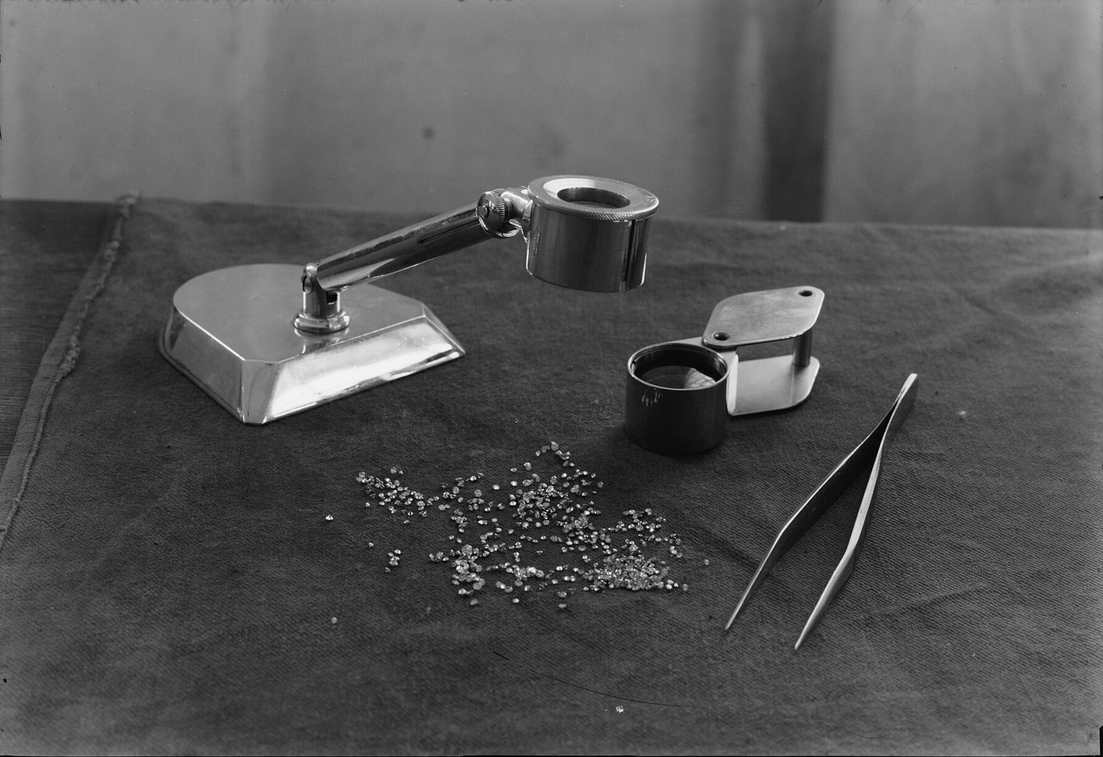 A jeweler's loupe stand, a folding loupe and tweezers beside a small parcel of loose polished diamonds laid out on cloth for inspection