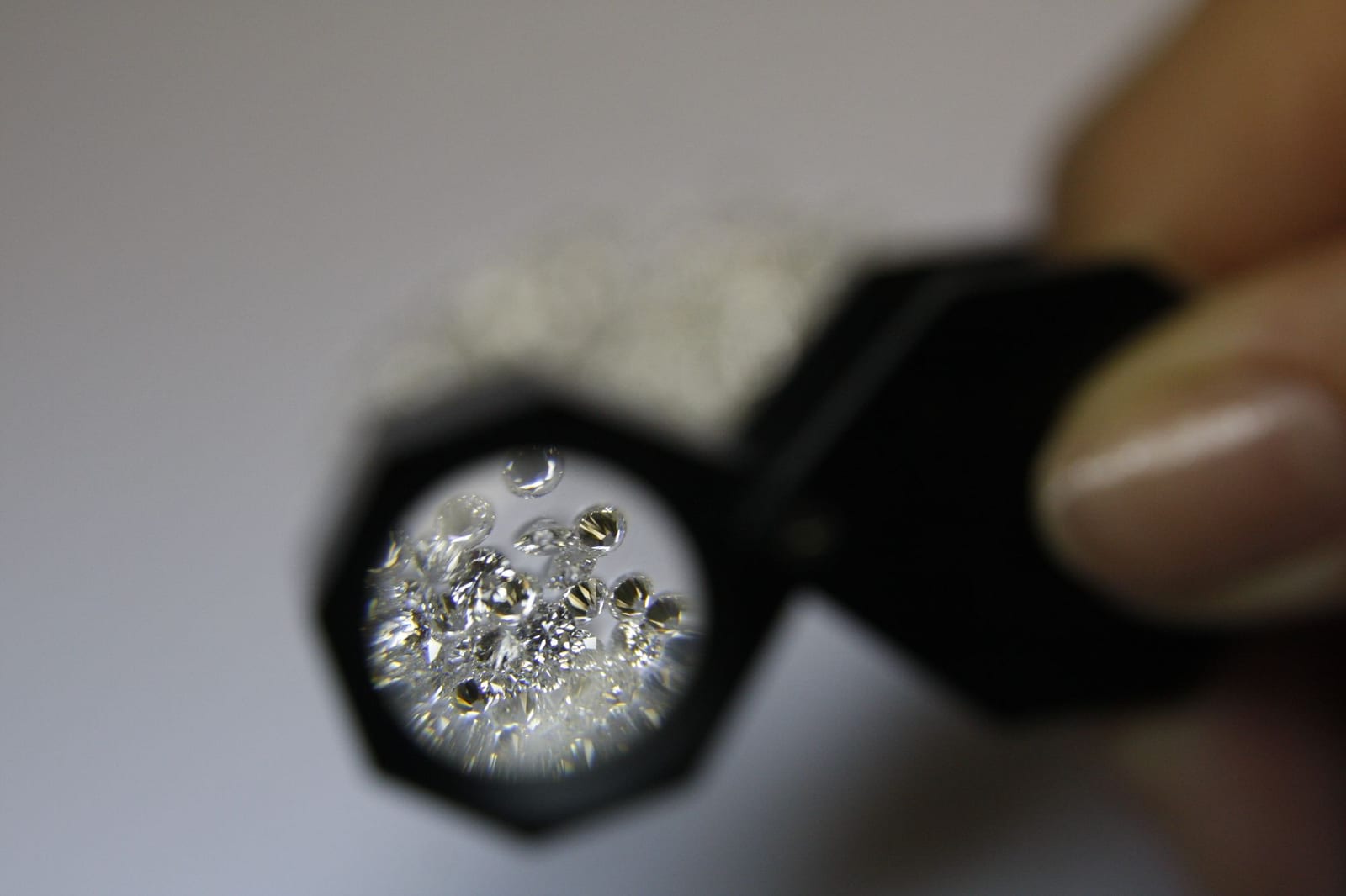 A folding jeweller's loupe held in the fingers over a small heap of round brilliant diamonds, the stones inside the lens magnified and sharp while the rest of the parcel sits blurred behind it