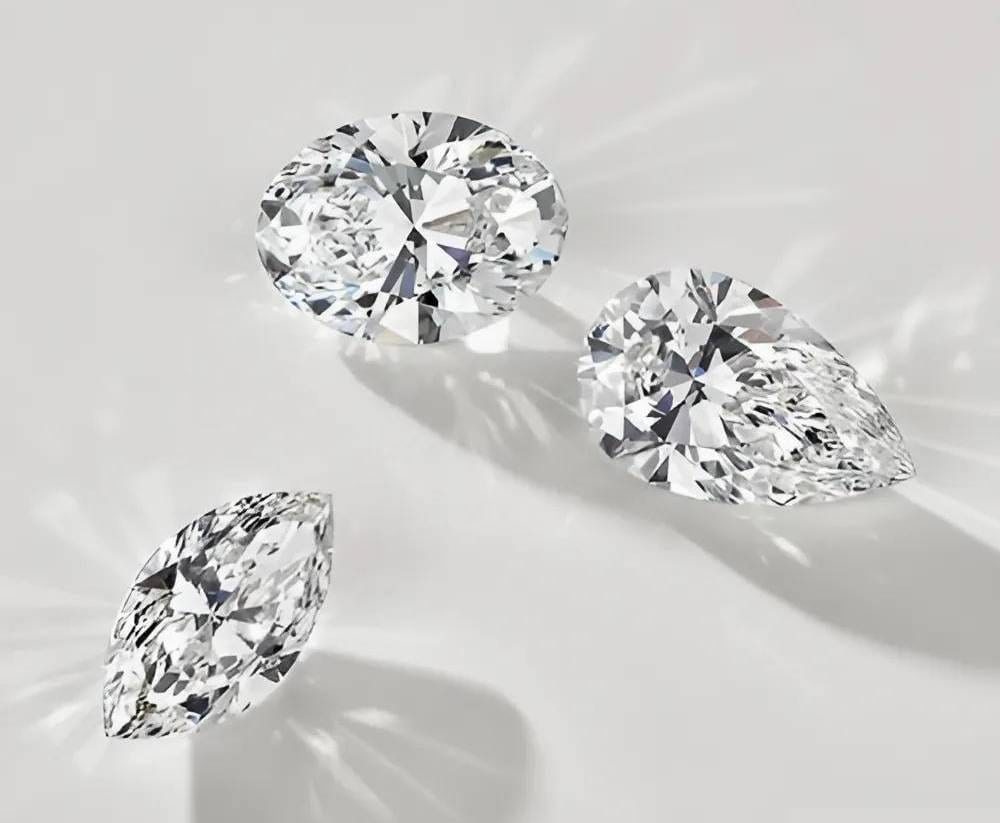 Three loose diamonds resting face up on a pale grey surface, an oval at the upper left, a pear at the right and a marquise below, each throwing a starburst of light across the background