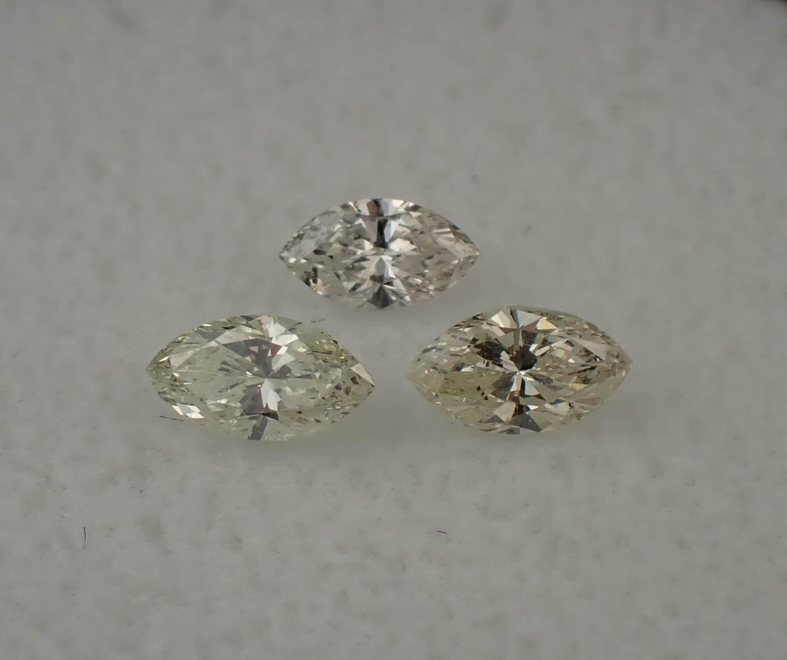 Three loose marquise cut diamonds arranged on a pale grey surface, one narrower than the other two, each showing a different body tint and a dark band running across the middle of the stone