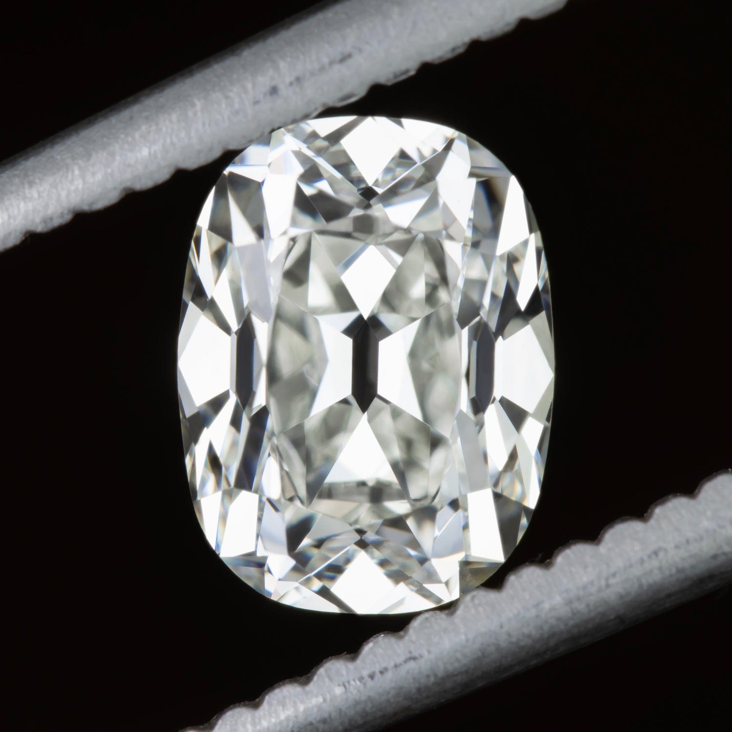 A loose cushion cut diamond held in fine tweezers against a plain black background, photographed face up. The outline is clearly taller than it is wide, with four softly rounded corners, nearly straight sides and broad chunky facets radiating from the centre.
