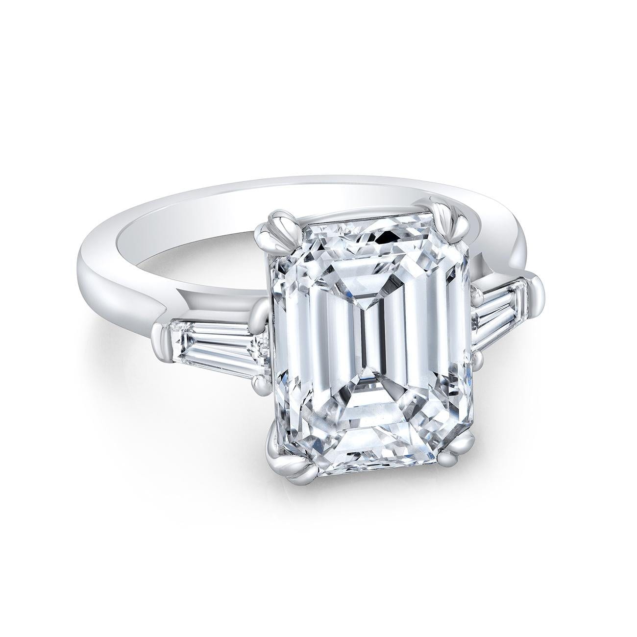 A three stone ring photographed at a slight angle on a plain white ground, a large rectangular emerald cut diamond held by four double prongs, with a tapered baguette running outward along each shoulder of the white metal band.
