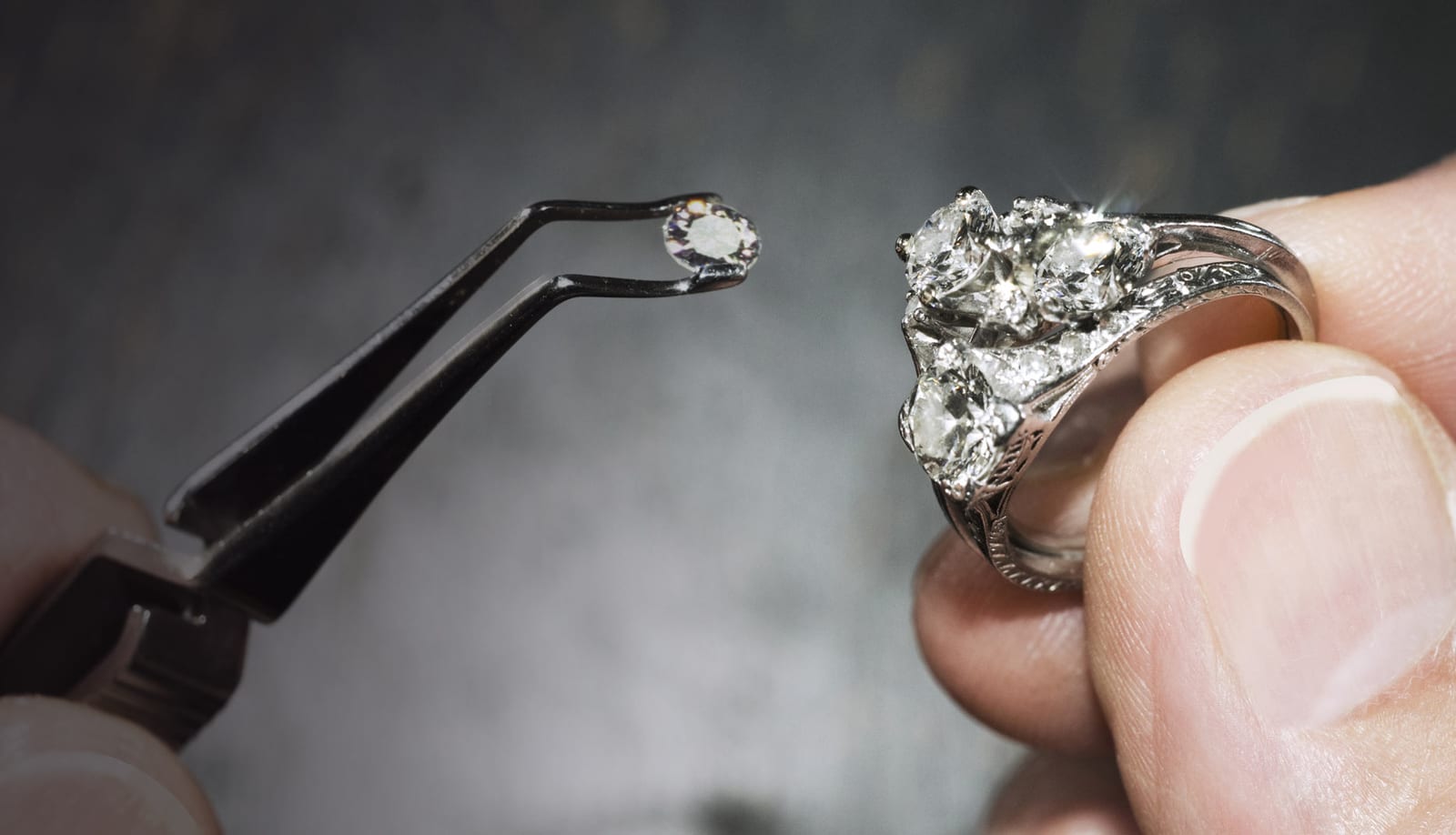 One hand holding a small loose round diamond in the jaws of black locking tweezers, raised level with a white metal diamond ring held in the other hand a few centimeters away.