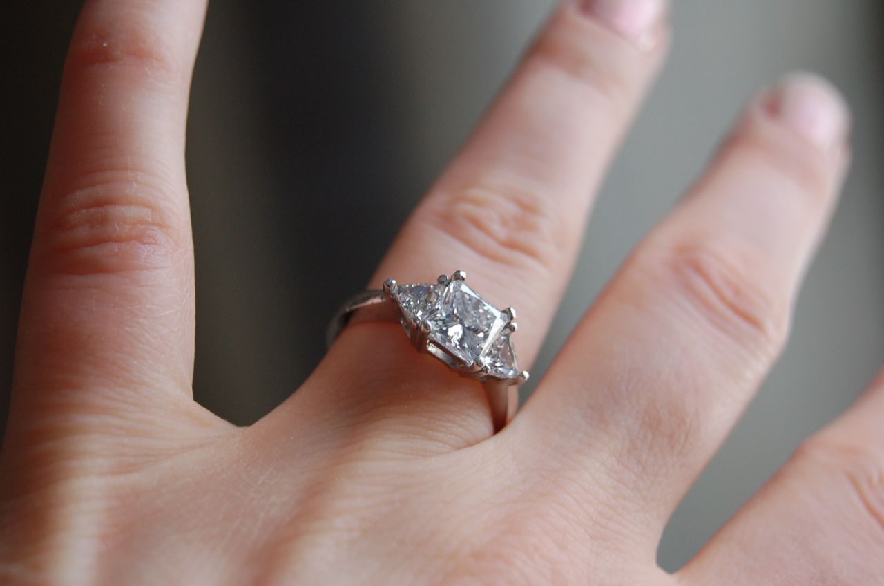 A platinum three stone engagement ring with a princess cut center diamond and two trillion cut side stones, worn on a hand