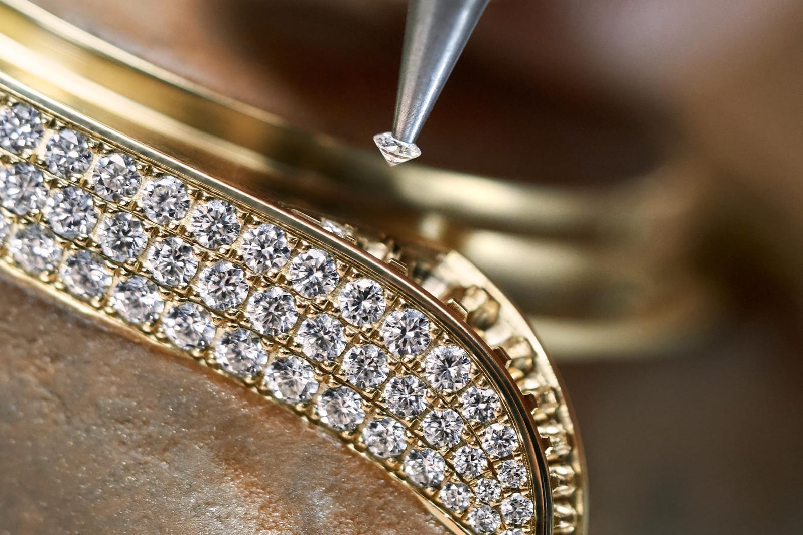 Close up of a curved yellow gold watch bezel set with tight rows of small round diamonds held by raised metal beads, with a pointed setting tool holding a single loose stone in the air just above the plain gold beside the rows.