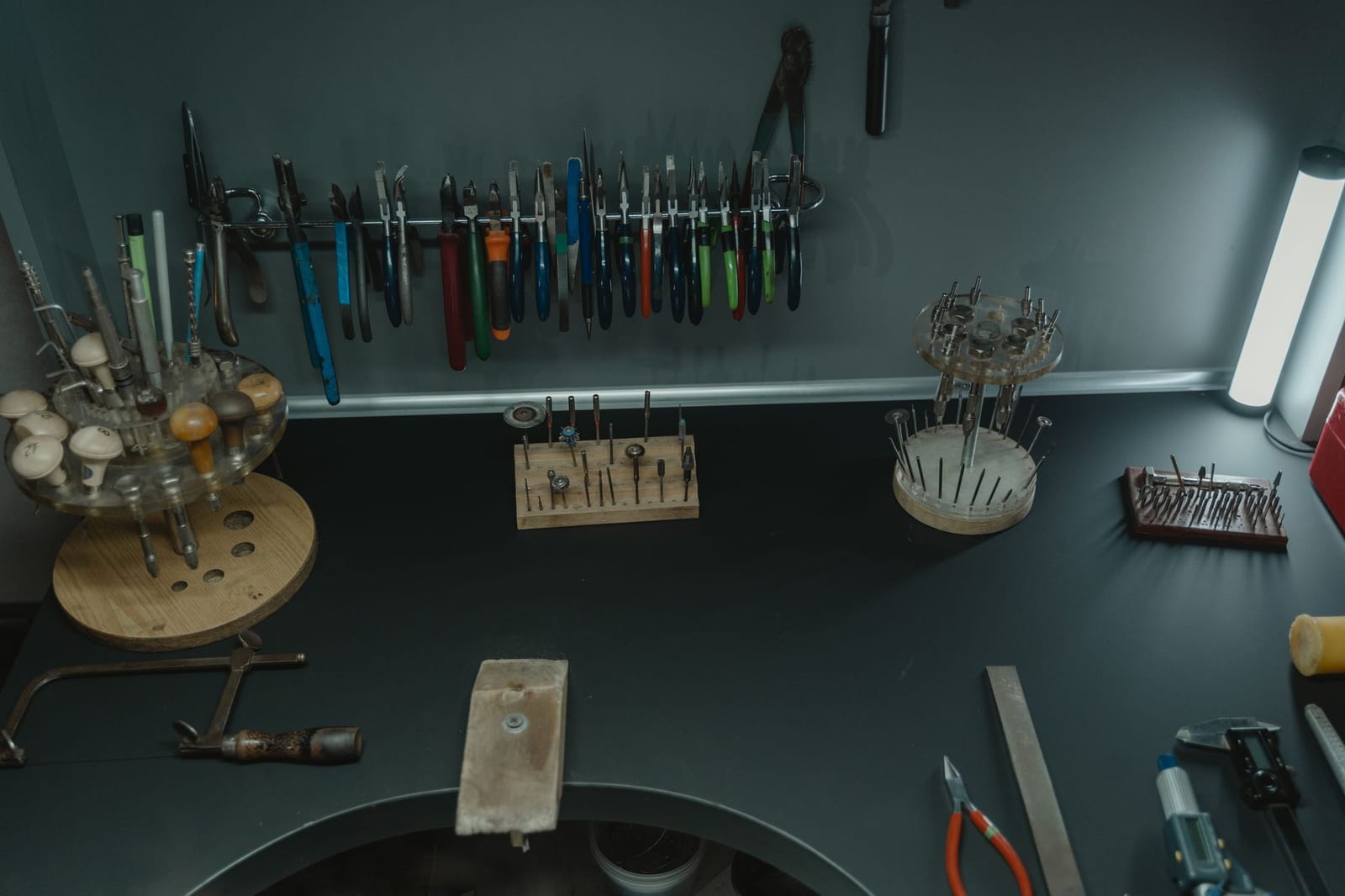 Overhead view of a jeweller's bench, with pliers and cutters hung in a row on a wall rail, wooden and acrylic stands holding burs and setting punches, a bench pin and a steel rule laid on the dark work surface