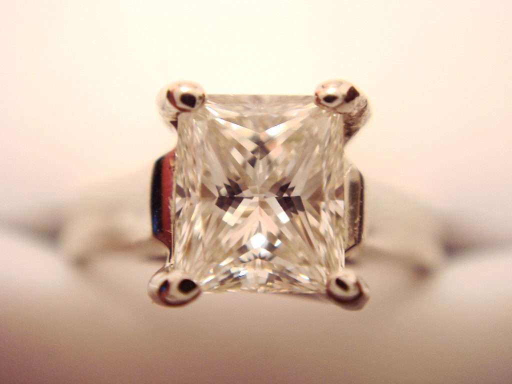 A close macro photograph of a princess cut diamond set in a four prong ring, showing the stone's facets and light return