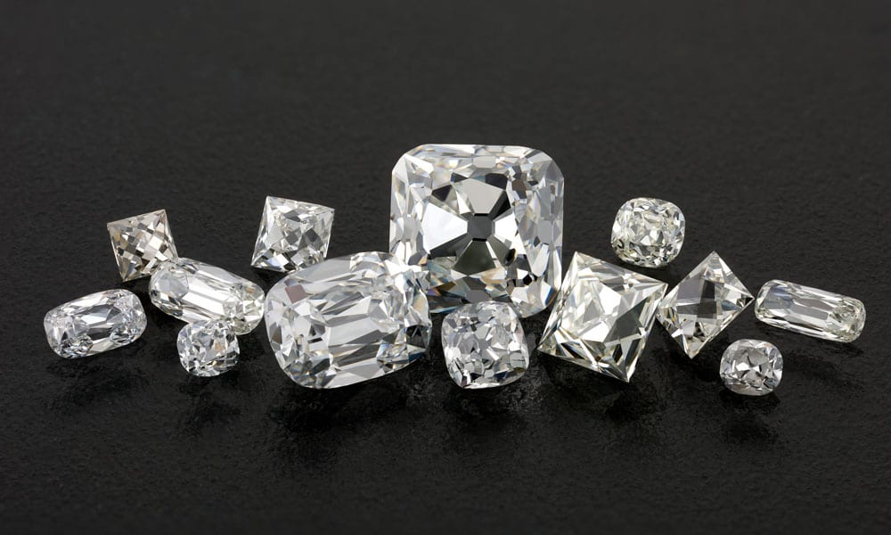 Thirteen or so loose antique cut diamonds heaped together on a black surface, ranging from a large squarish cushion in the middle to small stones at the edges, most of them chunky cushions with small tables and a dark culet showing at the center, with one long narrow step cut stone lying at the right edge