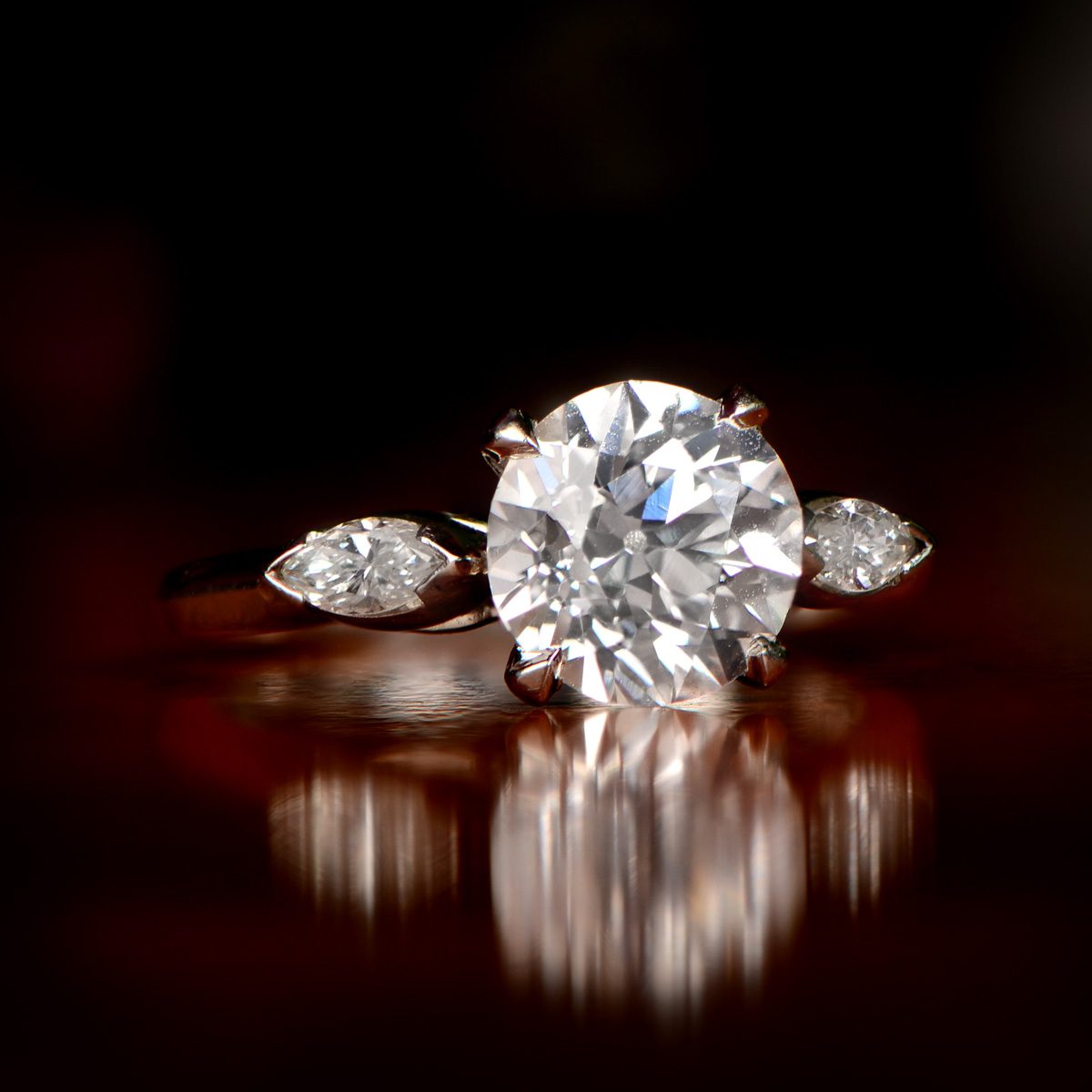 An antique diamond engagement ring from around 1920, the round stone showing the chunky faceting and high crown of a hand cut diamond, flanked by two small marquise side stones on a rose gold band