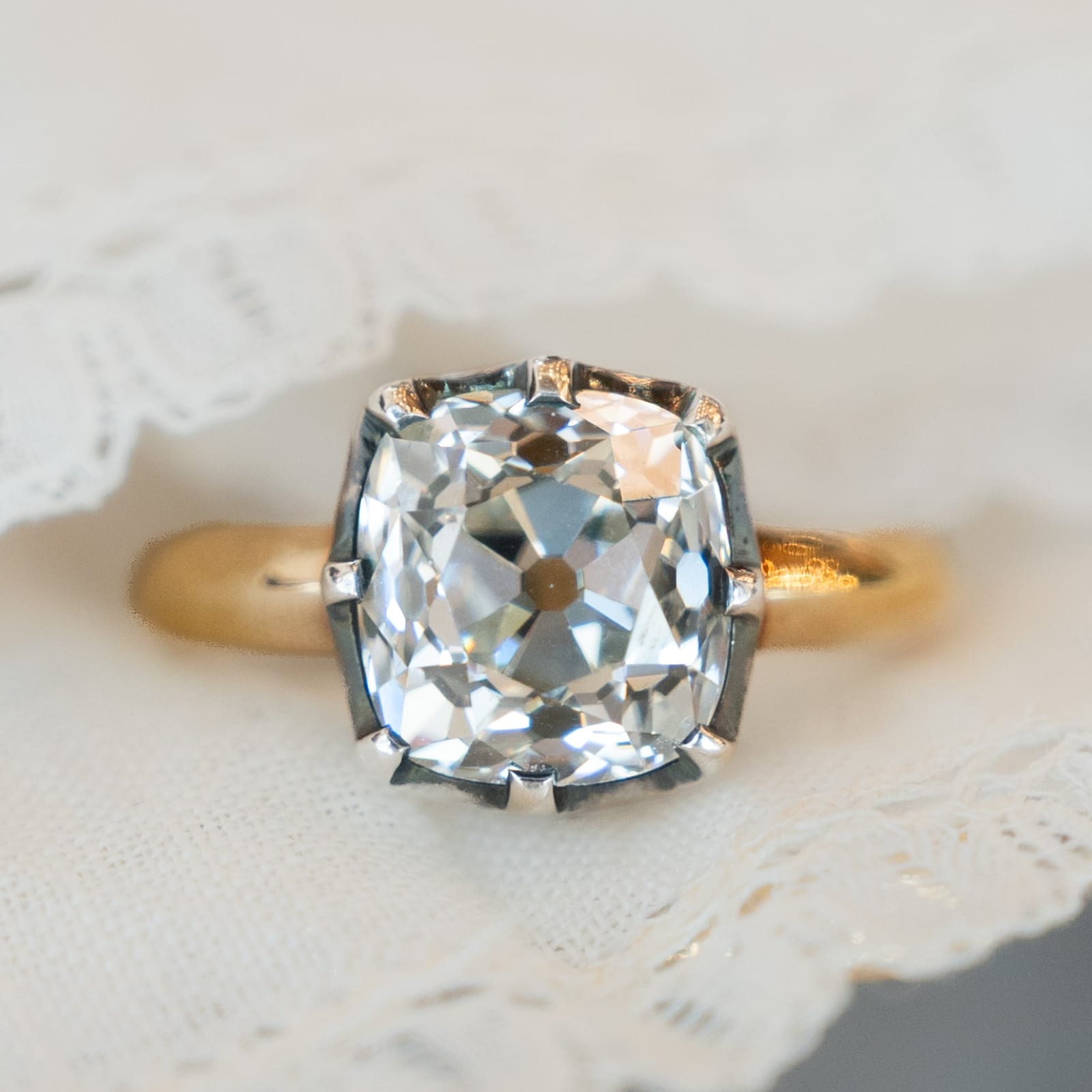 A soft squarish cushion outline diamond photographed straight down through its table, held in claws on a gold band resting on cream lace, with large uneven facets around the edge and a dark octagonal dot at the dead center of the stone
