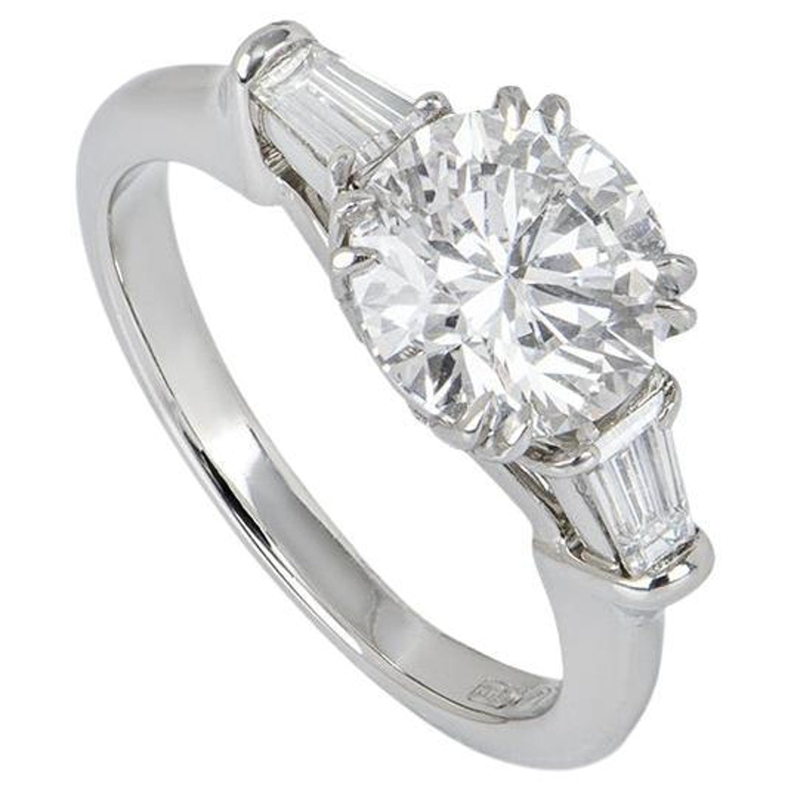 A platinum ring seen at a three quarter angle, a round brilliant held in prongs at the top and a tapered baguette set on each shoulder, the narrow end of each baguette running down into the polished band.